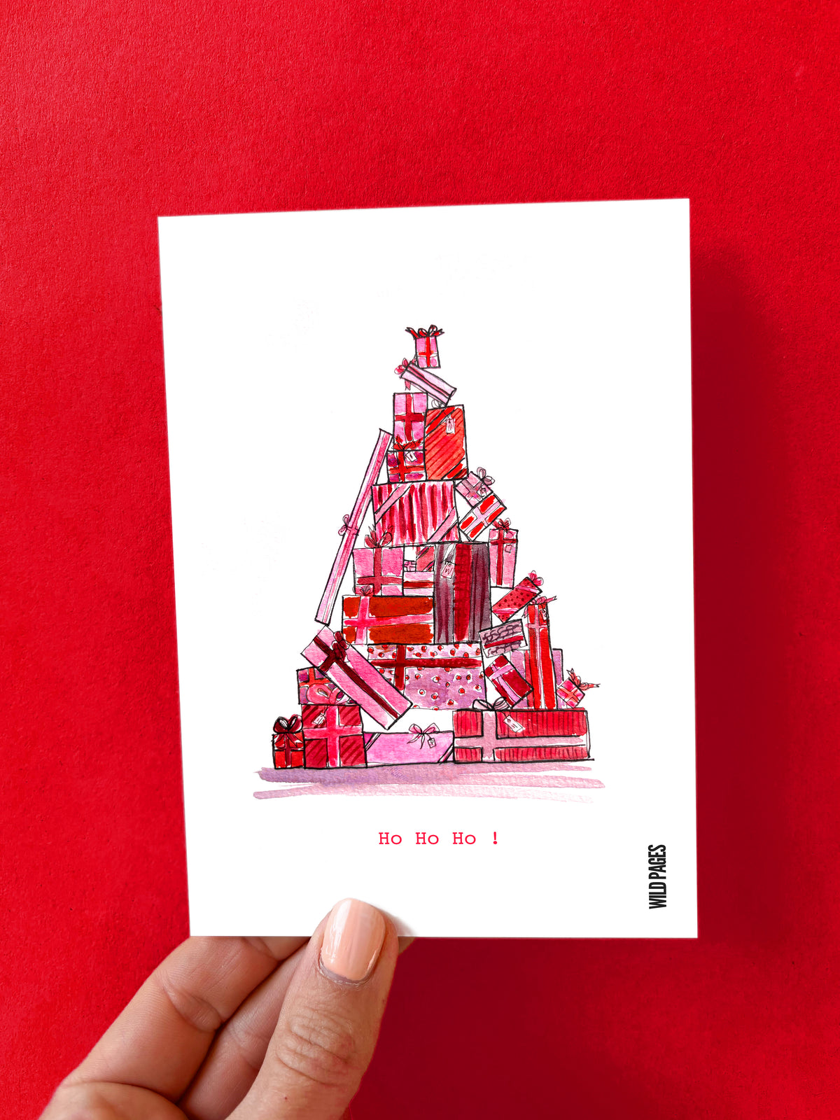 Illustration aquarelle d'une pyramide de cadeaux roses et rouges formant un sapin, texte « Ho Ho Ho ! », carte tenue devant un fond rouge.