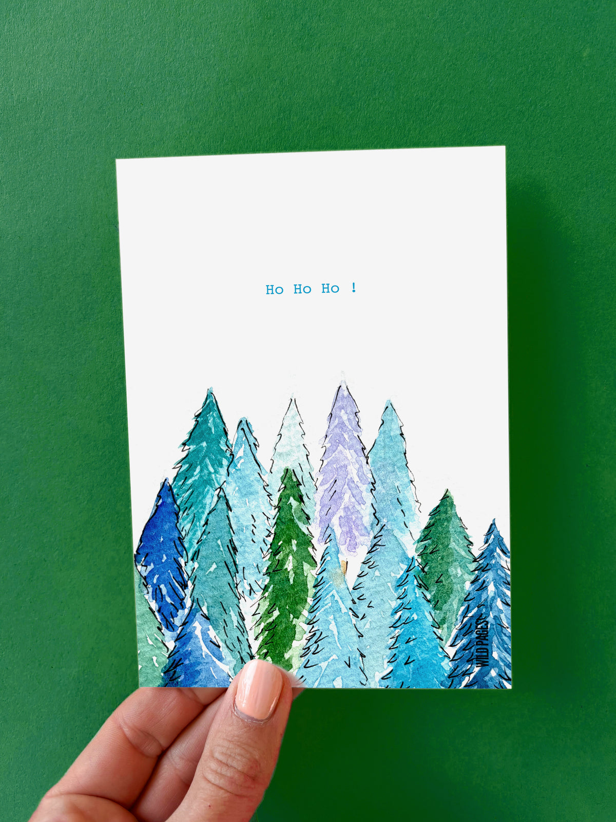 Illustration aquarelle d'une forêt de sapins en bleus et verts avec le texte « Ho Ho Ho ! », carte tenue devant un fond vert.