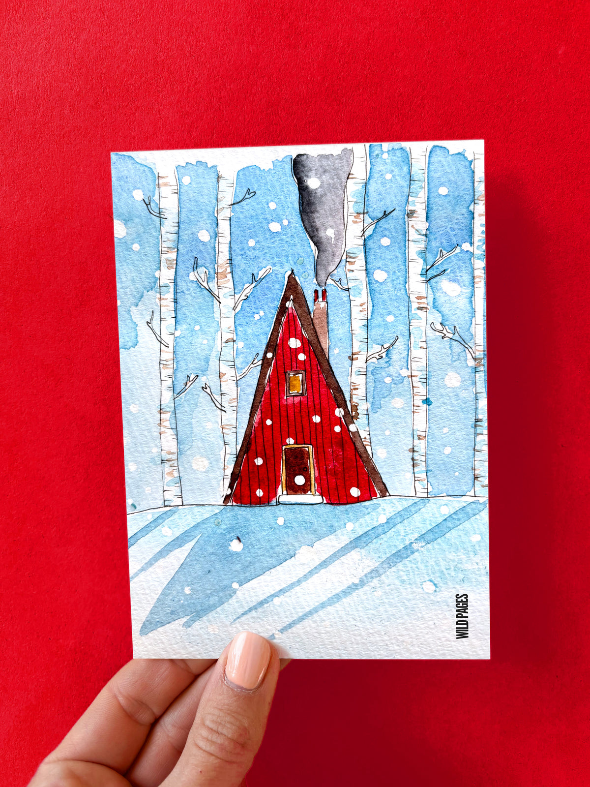 Illustration aquarelle d'un chalet rouge sous la neige au milieu de bouleaux blancs, carte tenue devant un fond rouge.