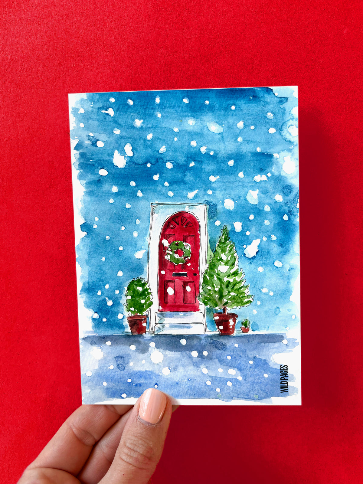 Illustration aquarelle d'une porte rouge avec une couronne de Noël, sous un ciel bleu enneigé, carte tenue devant un fond rouge.